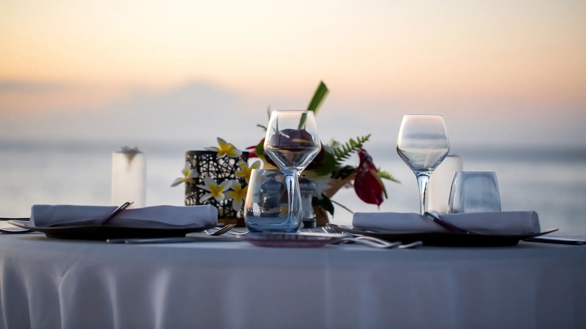 Elegantly set dinner table at a waterfront restaurant, featuring beautiful table settings with a blurred coastal view in the background.